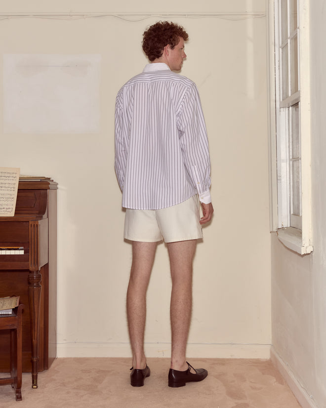 A man in a lightweight Steamboat Stripe Shirt with a contrasting white collar and white shorts stands facing a corner near a piano and window.