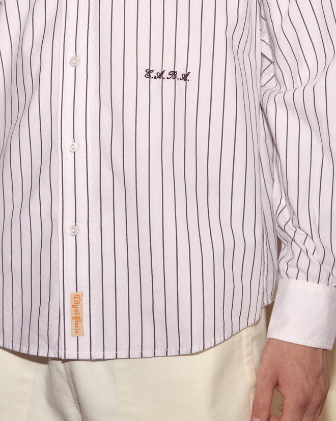 A person wears the Steamboat Stripe Shirt, a white pinstriped shirt with a contrasting white collar and embroidery, paired with cream pants and shown from the shoulders down.