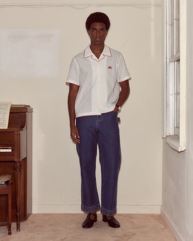 A person wearing a Redbud Short Sleeve Shirt and blue jeans stands indoors near a piano and a window.
