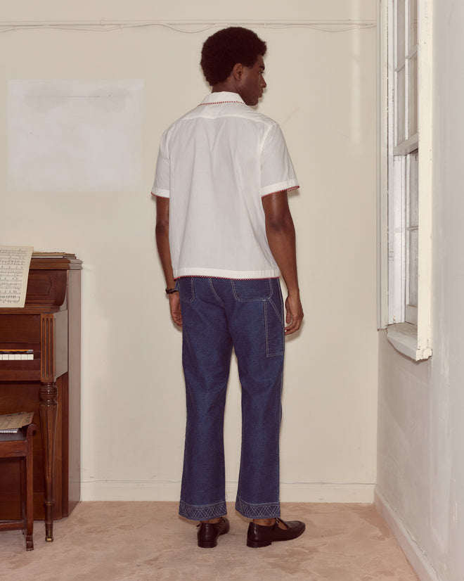 A person in a Redbud Short Sleeve Shirt and blue jeans stands with their back to the camera near a piano and window.
