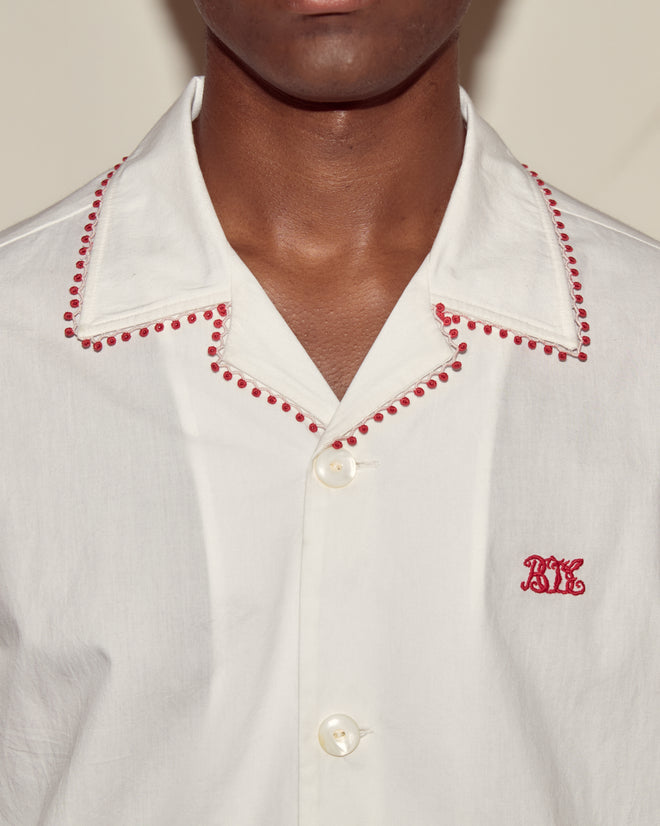 Man wearing the Redbud Short Sleeve Shirt, a white cotton voile shirt with red embroidery on the collar, red lace trim, and a Bode monogram on the chest.