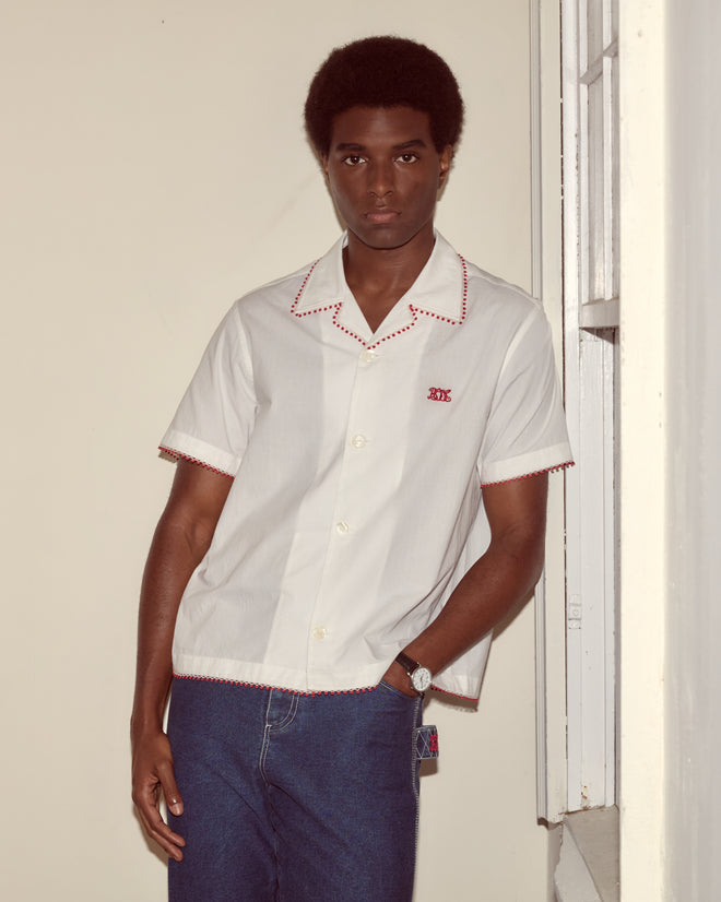 A man wearing the Redbud Short Sleeve Shirt and blue jeans stands by a cream-colored wall and window.
