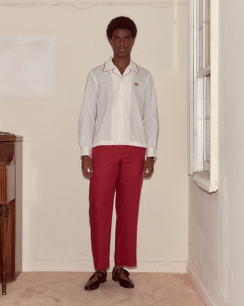 A man stands indoors near a window and piano, wearing a white Redbud Long Sleeve Shirt with a Bode monogram, red pants, and brown shoes.