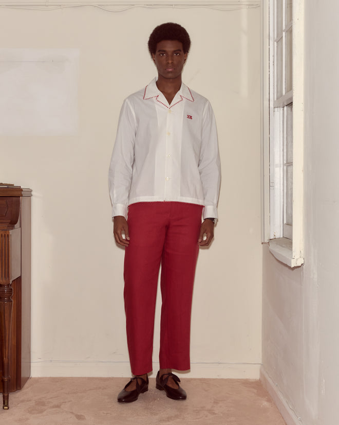 A man stands indoors near a window and piano, wearing a white Redbud Long Sleeve Shirt with a Bode monogram, red pants, and brown shoes.