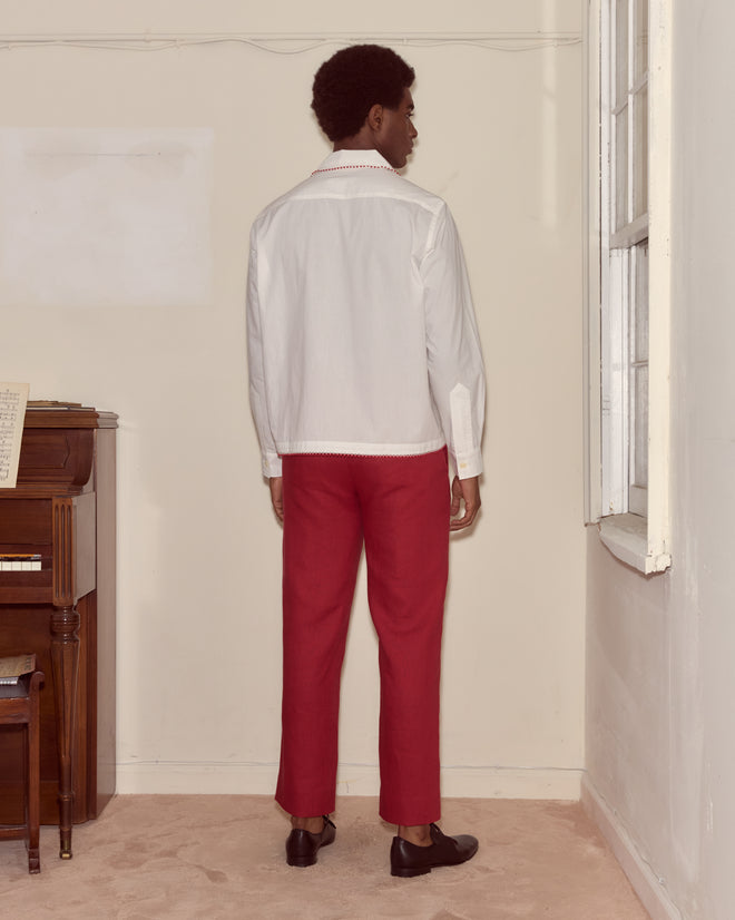 Indoors near a piano and window, a person faces away wearing the Redbud Long Sleeve Shirt—white cotton voile with red lace trim—paired with red pants and black shoes.
