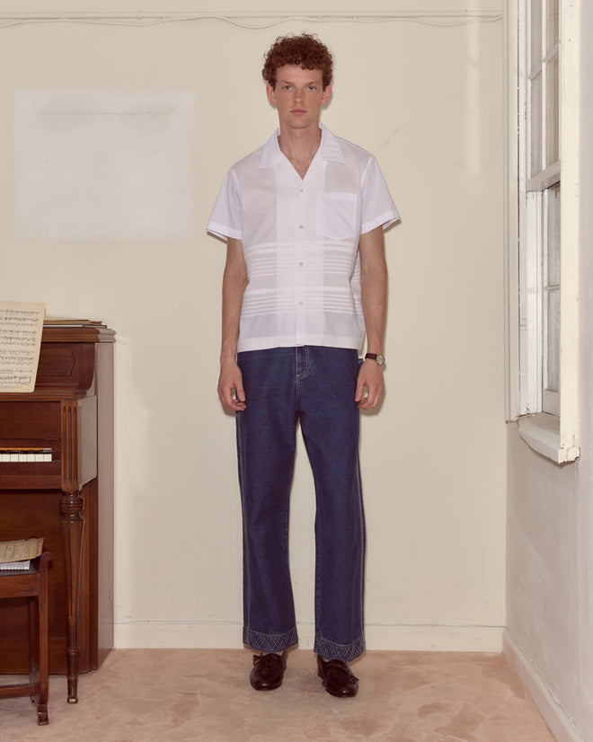 A man stands in a beige room near a piano, wearing the Chamomile Short Sleeve Shirt and blue jeans.