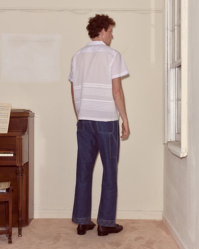 A man in the Chamomile Short Sleeve Shirt and jeans stands facing the corner of a room near a piano and a window.
