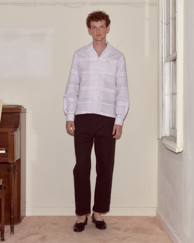 A man in a Chamomile Long Sleeve Shirt, black pants, and black shoes stands in a corner near a piano and a window.
