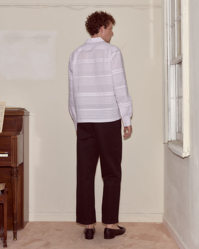 A man with curly hair stands facing a corner in a room, wearing the crisp white Chamomile Long Sleeve Shirt and black pants.