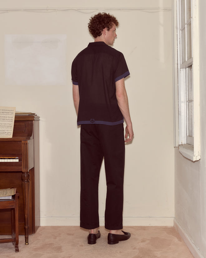 A person with curly hair wearing the Heart Deco Short Sleeve Shirt - Navy/Black stands facing a wall near a piano and window.