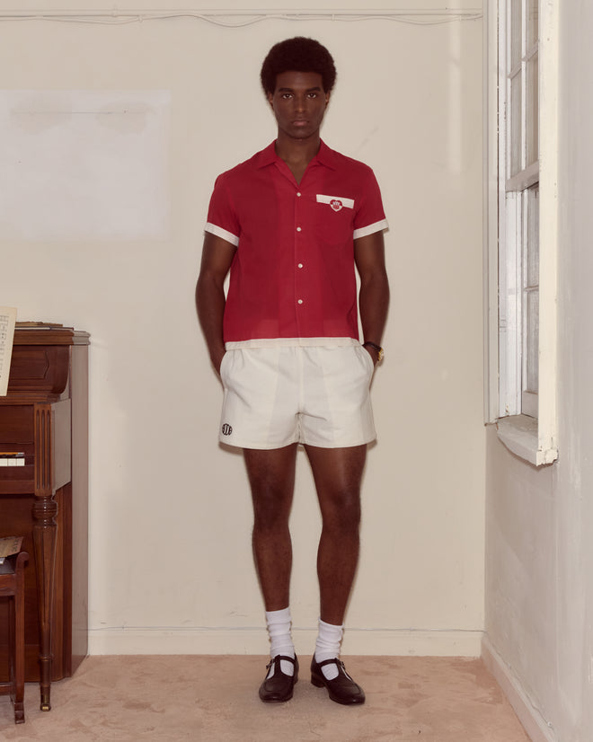 A man wearing the Heart Deco Short Sleeve Shirt in red/white and white shorts stands indoors by a piano and window, looking at the camera.
