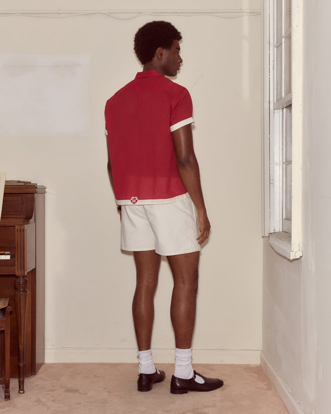 A person in a red and white Heart Deco Short Sleeve Shirt and white shorts stands facing a wall in a minimal room with a piano and window.