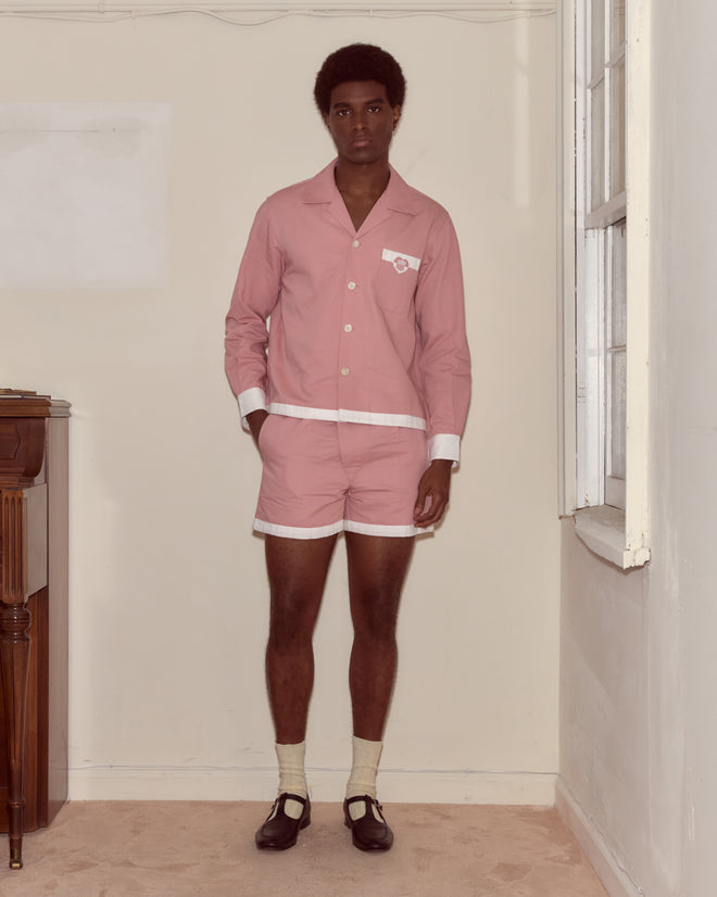 A person stands indoors wearing the Heart Deco Long Sleeve Shirt—cozy and 100% cotton—paired with pink pajamas with white trim, socks, and black shoes, looking at the camera.