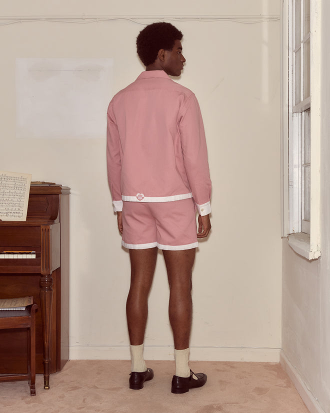 A man in pink shorts and a Heart Deco Long Sleeve Shirt stands facing a wall near a piano with sheet music on top.