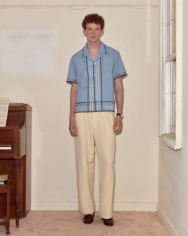 A man in a Ribbon Plaid Short Sleeve Shirt and cream pants stands indoors beside a piano, looking at the camera.