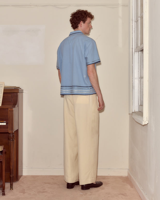 A man wearing a Ribbon Plaid Short Sleeve Shirt and cream trousers stands by the corner of a plain room near a window.