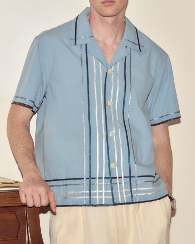 Man in a Ribbon Plaid Short Sleeve Shirt, standing by a wooden table.