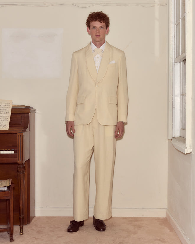 A man in a Slub Weave Tuxedo Jacket with a shawl collar stands indoors by a piano and window, looking directly at the camera.