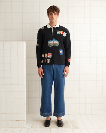 A person wearing a Multi-Patch Rugby Shirt in black and blue jeans stands in a tiled room.