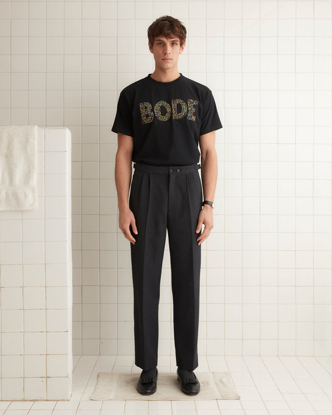 A person is standing straight against a beige wall, wearing the Multi Bead Tee by BODE, black pleated pants, and black shoes.