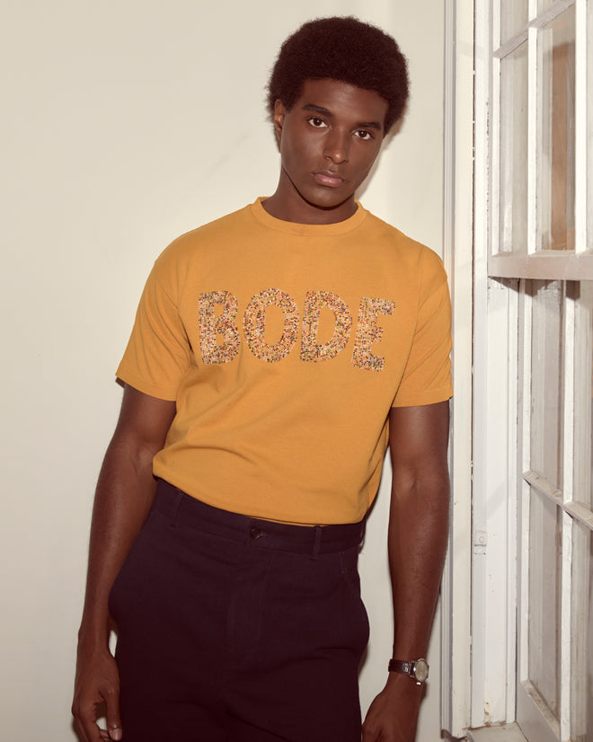 A man in a yellow "Multi Bead Tee" stands indoors by a window, looking at the camera.