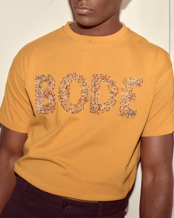 Person wearing a yellow Multi Bead Tee, made from 100% cotton and featuring “BODE” in patterned letters on the front, resembling a stylish beaded Bode T-shirt.