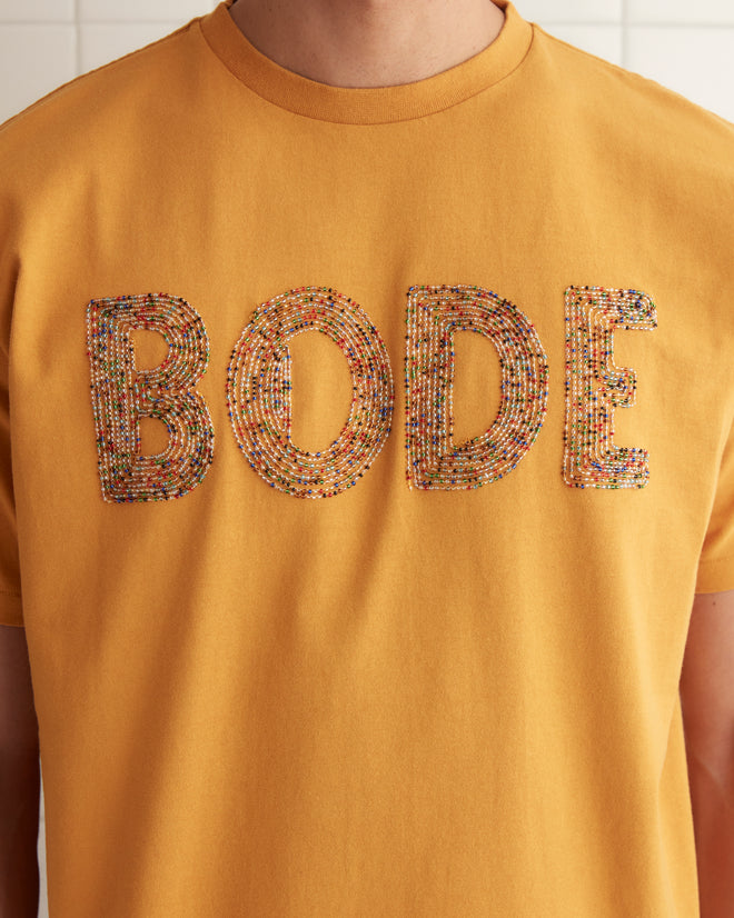 A person stands indoors against a plain wall, sporting the Multi Bead Tee—a yellow children’s novelty t-shirt with the word 