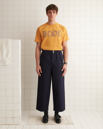 A person stands indoors against a plain wall, sporting the Multi Bead Tee—a yellow children’s novelty t-shirt with the word 