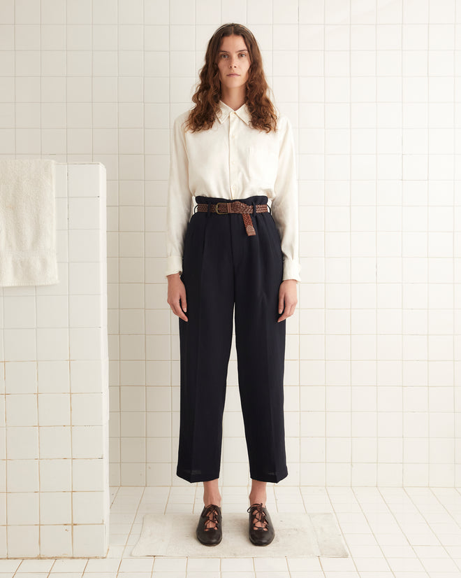 A person stands in a tiled room wearing a white shirt, the Murray Trousers - Midnight with a belt, and brown shoes.