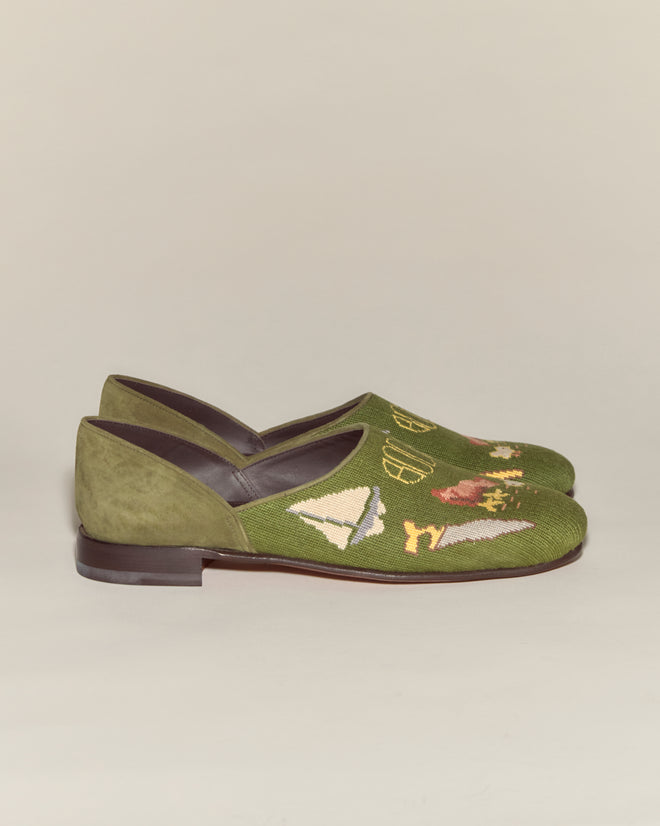 The Needlepoint House Shoes are green slip-ons with brown soles, inspired by the classic Bode House Shoe and made in Italy. They feature embroidered sailing and countryside scenes on the toe.