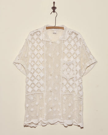 The Netted Flower Shirt - S, a unique short-sleeve white crochet lace top, is displayed on a wooden hanger against a plain wall.