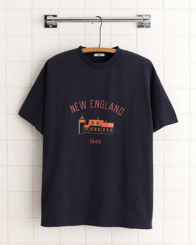 The New England Tee - Midnight, featuring "New England 1945" and a building graphic screen-printed on the front, is displayed on a white tiled wall.