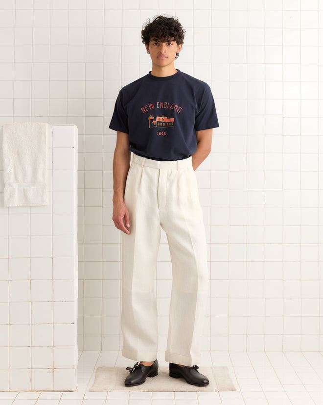 A man stands in a tiled room wearing the New England Tee - Midnight, white trousers, and black shoes.