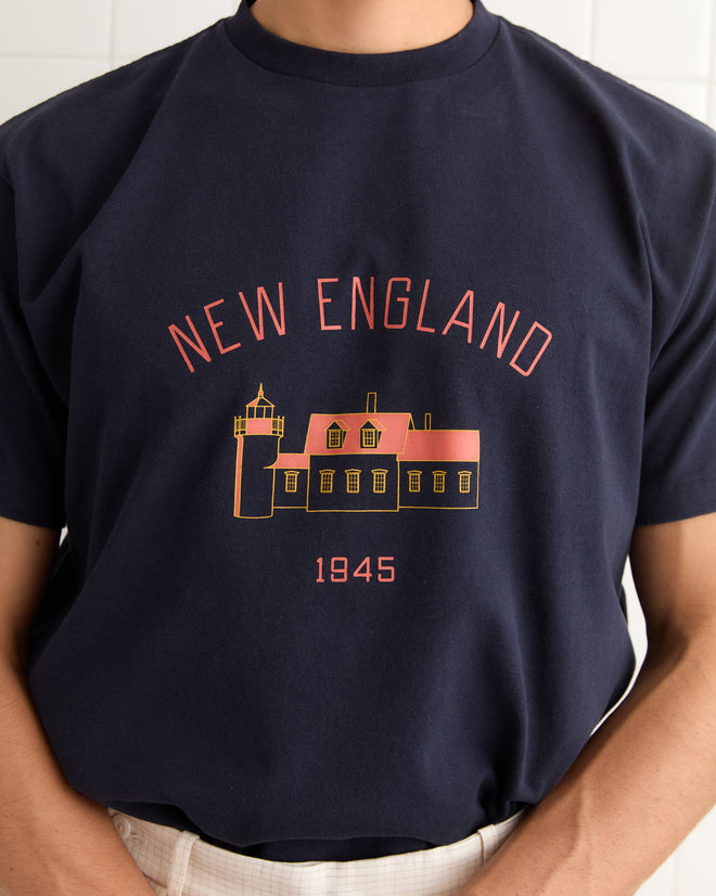 Person wearing the New England Tee - Midnight, a navy short sleeve shirt featuring a "NEW ENGLAND 1945" print with a lighthouse and house graphic on the front.
