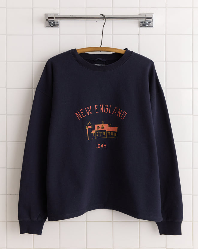 The New England Crewneck - Midnight is a navy blue 100% cotton sweatshirt featuring "NEW ENGLAND 1945" and a lighthouse graphic, inspired by classic souvenir designs, shown on a wooden hanger.
