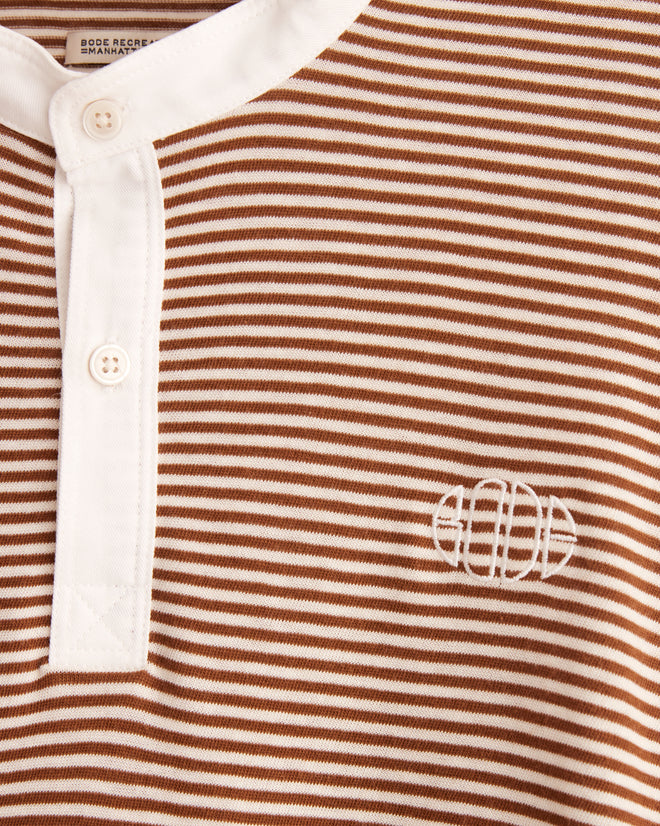 The Newton Stripe Polo, featuring brown and white stripes with a classic style and a crisp white collar, is displayed on a hanger against a tiled wall.