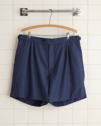 The Night Blue Wool Shorts - 34, crafted from men's suiting fabric for a perfect blend of style and durability, hang on a wooden hanger against a white tiled wall.