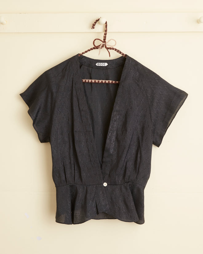 The Night Tide Top - 10, a unique black short-sleeve blouse, elegantly hangs on a hanger against a beige wall, capturing the timeless charm of the early 20th century.