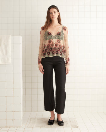 A woman wearing the Solarium Top, featuring a floral print and a swing silhouette, paired with black pants stands on a tiled floor in front of a white wall.