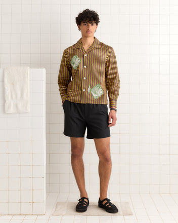 A man wearing a Fish Appliqué Long Sleeve Shirt and black shorts stands in a white tiled room beside a towel on a partition.