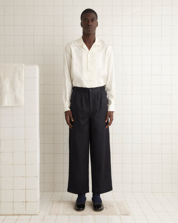 A person wearing Numerals Trousers and a white shirt stands in a tiled room next to a towel.