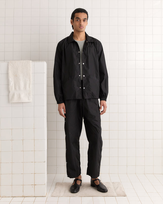 A person in a Nylon Track Jacket - Black stands in a tiled room, with a nearby towel hanging.