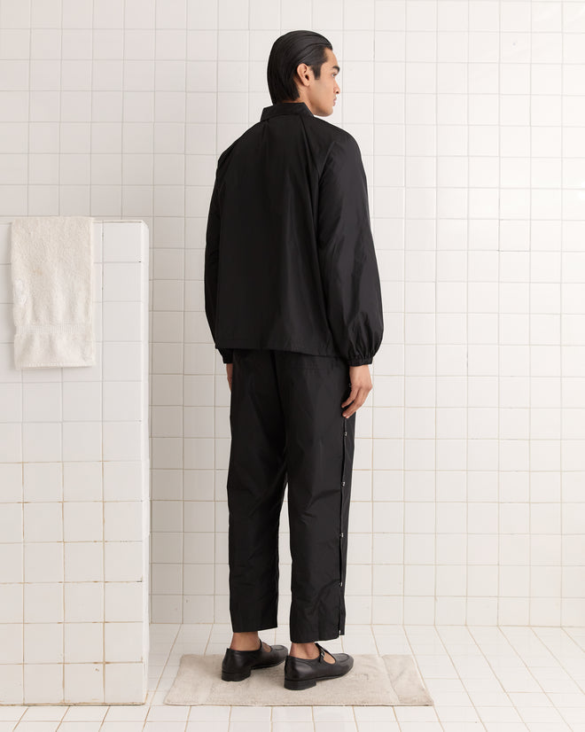 A person in a black Nylon Track Jacket stands on a rug in a white tiled bathroom, facing the wall, with enamel horseshoe-and-star snap buttons catching the light.