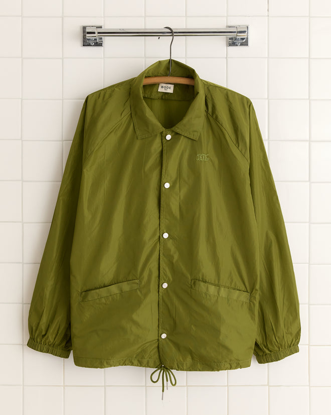 The Nylon Track Jacket in olive, featuring enamel snap buttons, hangs on a tiled wall.