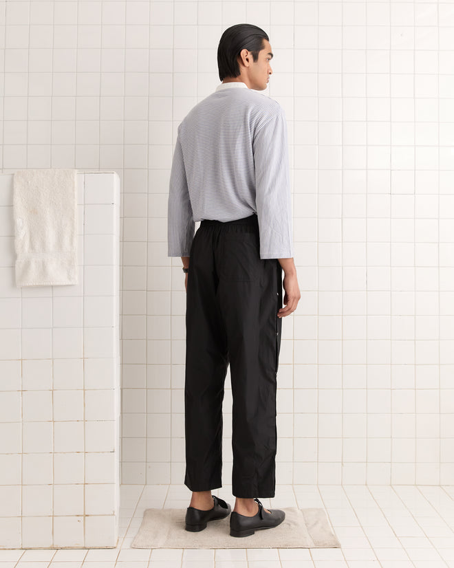 A person stands in a tiled room, wearing a gray top and trendy Nylon Tear-Away Track Pants - Black, facing a wall with a hanging towel.