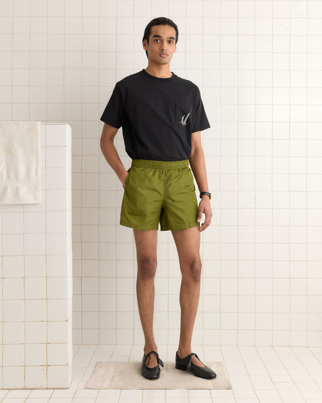 A person in a black shirt and olive Nylon Track Shorts with "Bode" embroidery stands in a tiled room, a folded towel visible nearby.