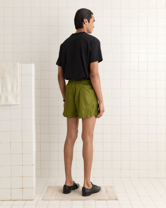 A person in a black shirt and olive Nylon Track Shorts featuring subtle "Bode" embroidery stands on a bath mat in a white-tiled room, facing away.