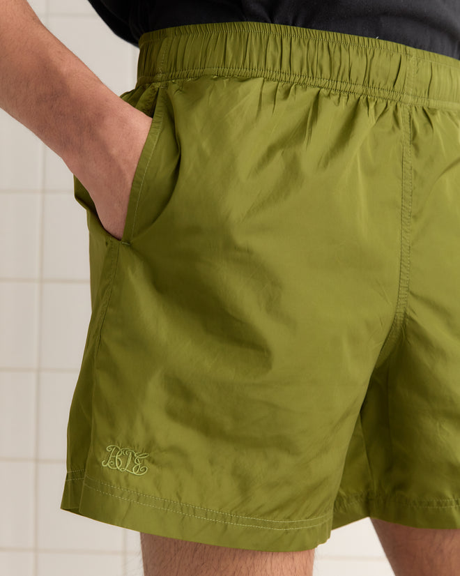 In olive Nylon Track Shorts, beautifully crafted from lightweight fabric with "Bode" embroidery, a person stands casually against a tiled background, one hand resting in the pocket.