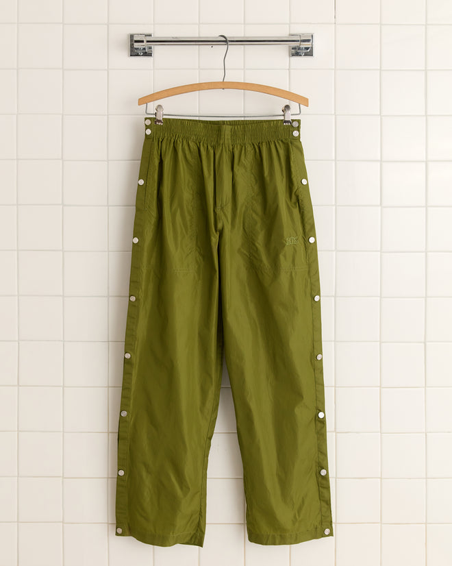 Nylon Tear-Away Track Pants in olive, reminiscent of 90s tearaway style, hang on a wooden hanger against a tiled wall.