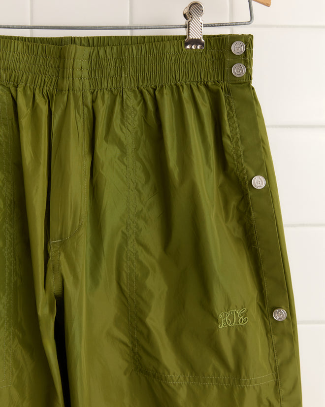 Olive Nylon Tear-Away Track Pants with a drawstring waist and decorative side buttons, invoking a subtle 1990s style, displayed on a hanger.